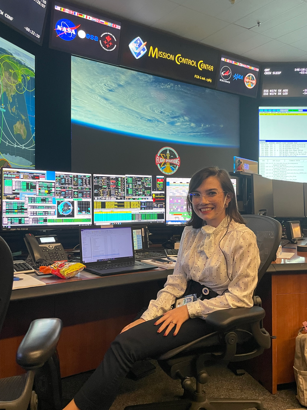 A woman smiling and sitting in a chair at a desk with a computer, three monitors, and a large screen with a picture of Earth from space behind her. 